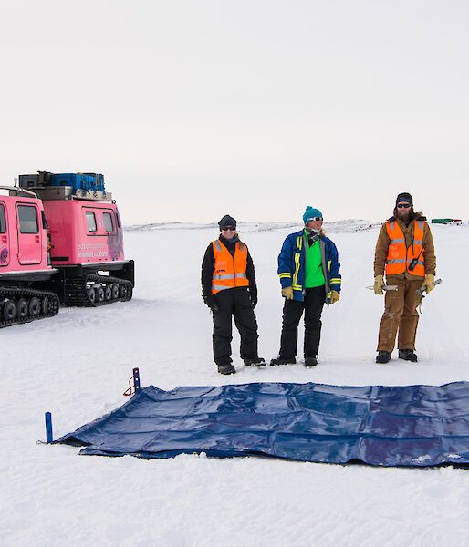 Expeditioners standing near marked out area on sea ice with snow pole line marker and flag with oversnow vehicle behind