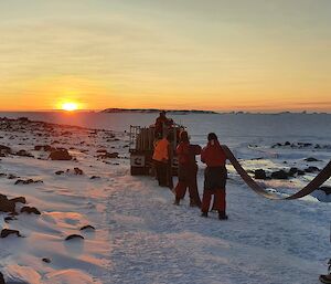 Expeditioners holding a flat fire hose over their shoulders against an orange sunset.