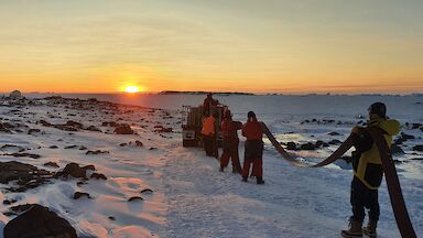 Expeditioners holding a flat fire hose over their shoulders against an orange sunset.