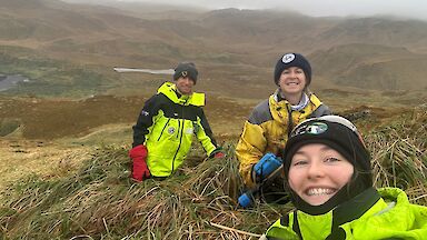 Three smiling people wearing yellow outdoor clothing stand in a misty, hilly landscape covered in tussock.