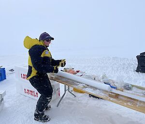 An expeditioner stands next to a long ice core on a table in an icy landscape.