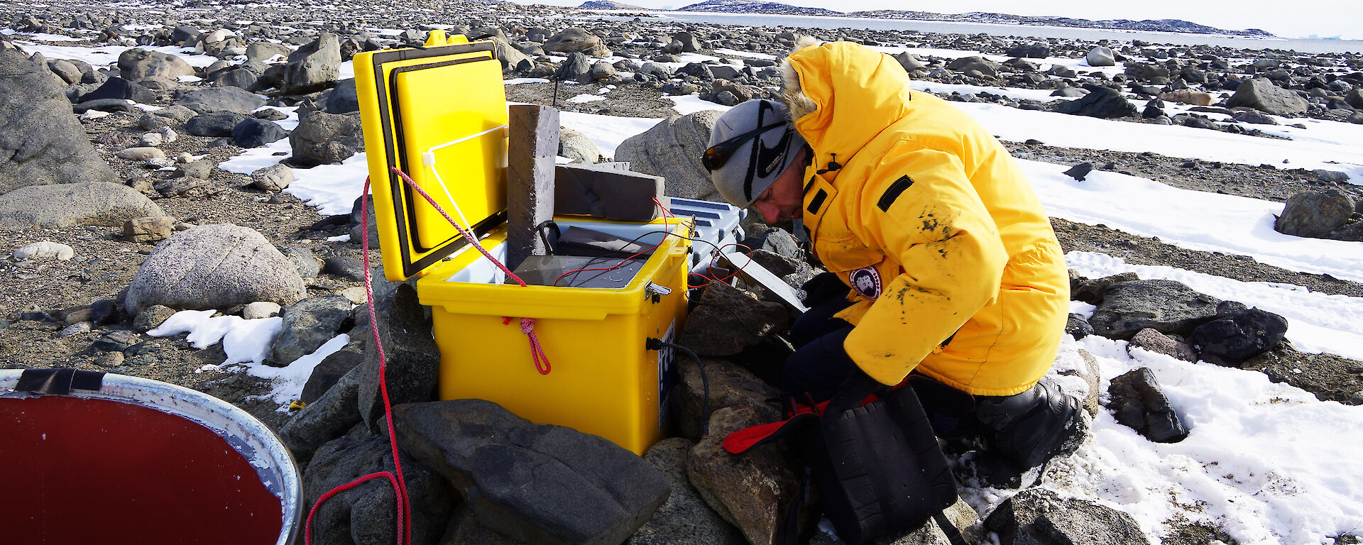 Man kneeling in front of yellow plastic box adjusting electronics equipment, on snow and rock covered ground.