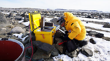 Man kneeling in front of yellow plastic box adjusting electronics equipment, on snow and rock covered ground.